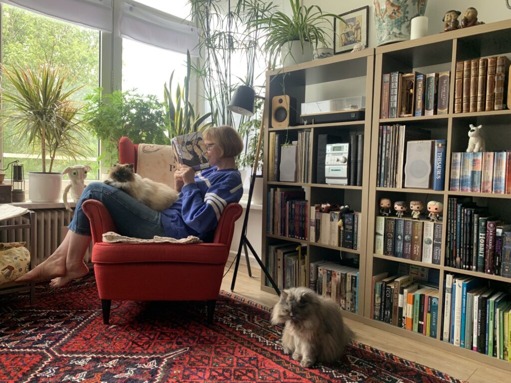 Astrid (een witte vrouw) zit op een rode stoel, met haar benen over de leuning. Ze houdt een boek dicht bij haar gezicht. Op schoot en op de grond voor de stoel een pluizige grijze kat.