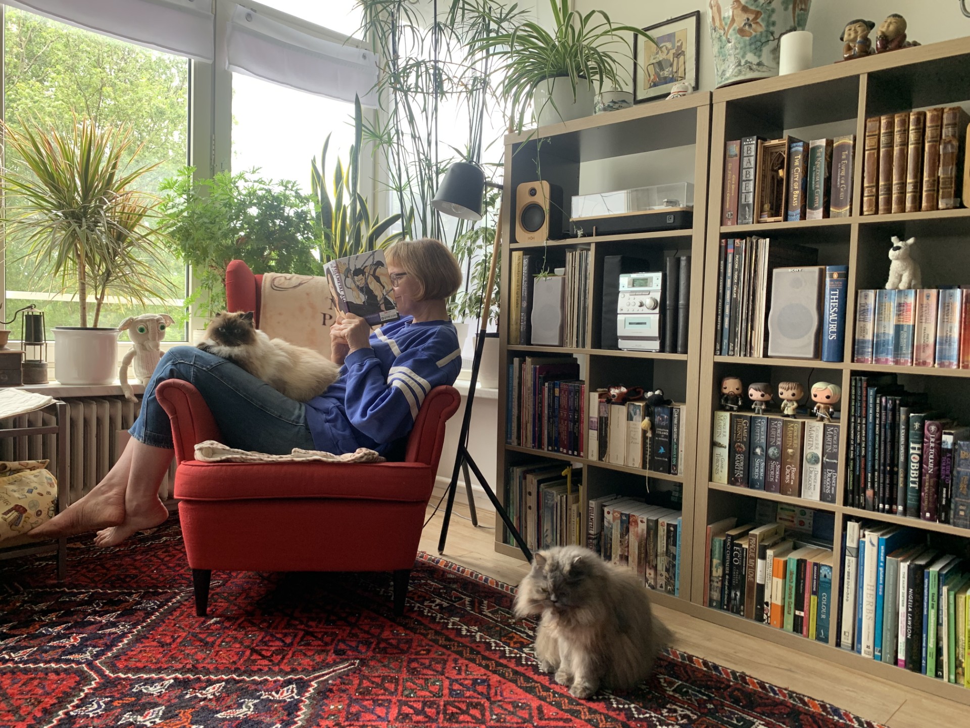 Astrid (een witte vrouw) zit op een rode stoel, met haar benen over de leuning. Ze houdt een boek dicht bij haar gezicht. Op schoot en op de grond voor de stoel een pluizige grijze kat.