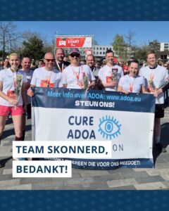 Team Skonnerd poseert met Cure ADOA Foundation banner bij de finish met medailles om hun nek en bloemen in hun handen.