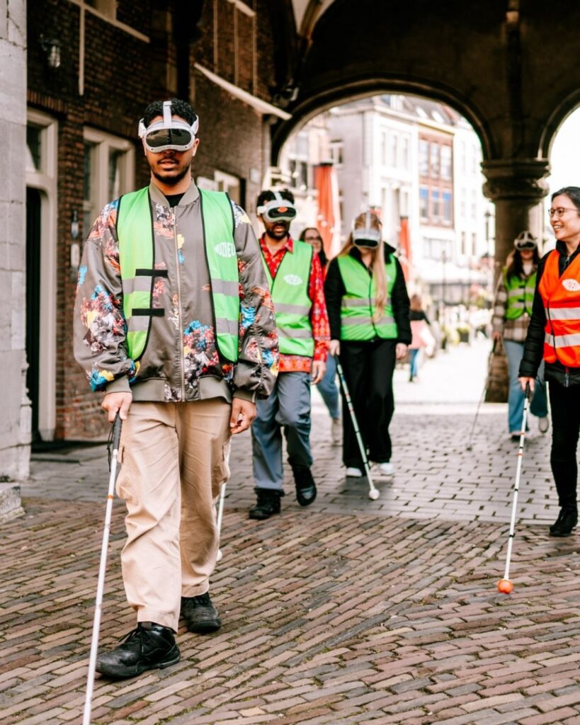 Groep muZIEum bezoekers loopt met VR-brillen en taststokken door stad onder begeleiding van slechtziende gids.