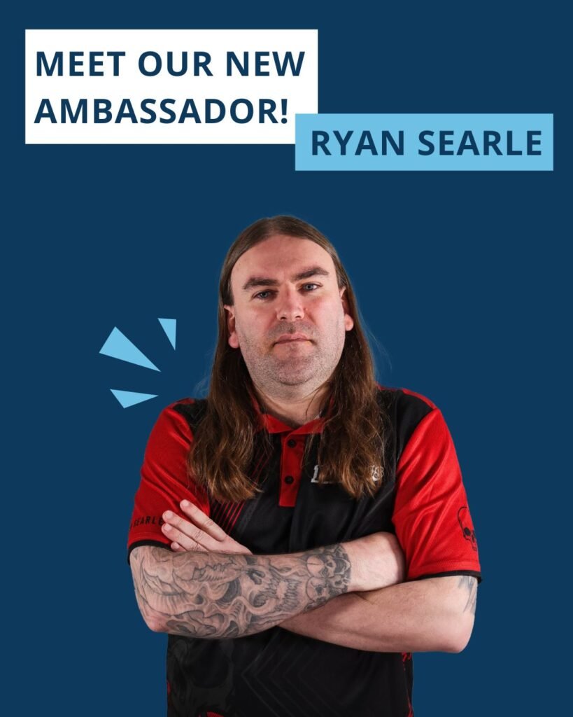 Een foto van professioneel darter Ryan Searle in actie, gekleed in een zwart-rood shirt met een afbeelding van een skelet. De afbeelding bevat de tekst ‘MEET OUR NEW AMBASSADOR!’ en ‘RYAN SEARLE’