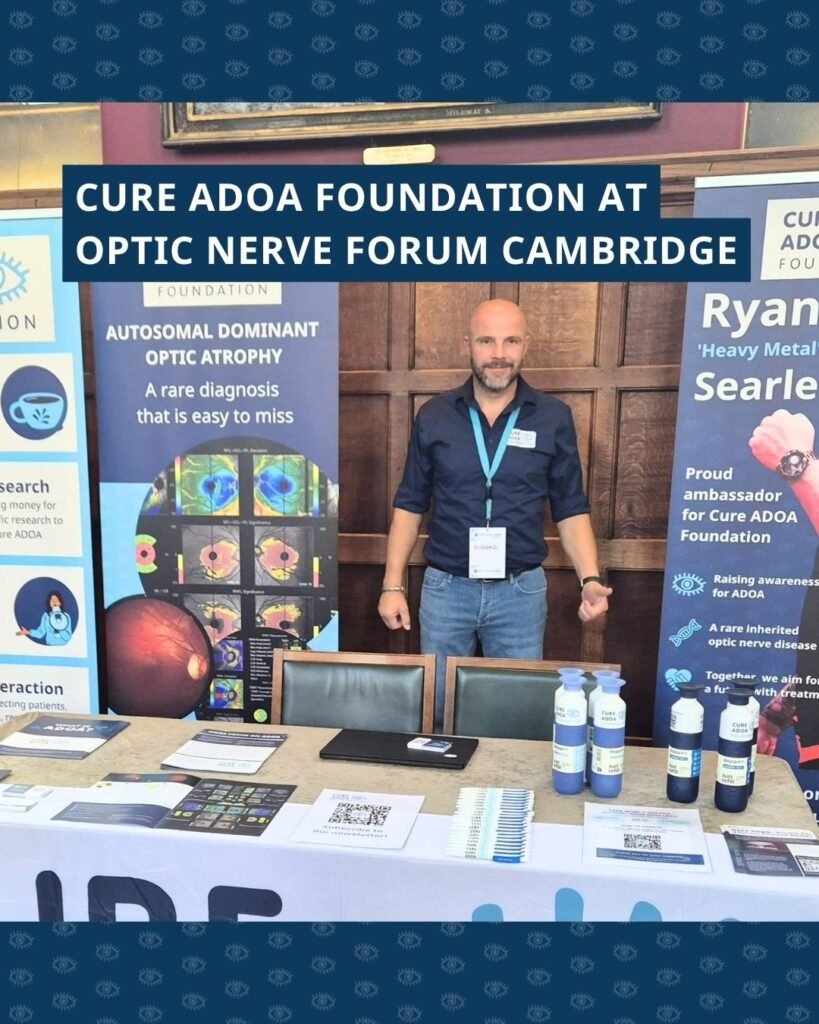 Richard bij de stand van de Cure ADOA Foundation. Op tafel ligt voorlichtings- en promotiemateriaal. Naast hem staan verschillende banners. Bovenin de foto staat de tekst ‘Cure ADOA Foundation at Optic Nerve Forum Cambridge’.